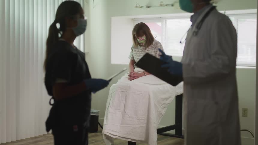 Male doctor and female nurse discuss female patients medical records as the patient sits on the examination table with her head down