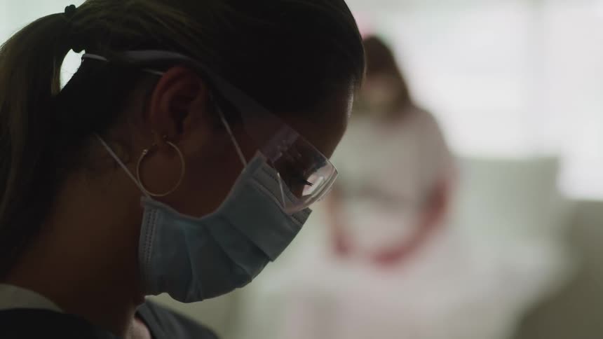 A woman with scrubs a mask and goggles looking down as another woman sits in the background