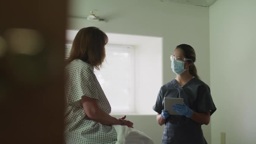 Female nurse wearing latex gloves and holding a tablet has a conversation with a female sitting on an examination table wearing a patient gown and face mask