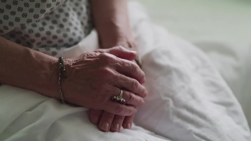 Elderly woman sits with a sheet on her lap as she clasps her hands together