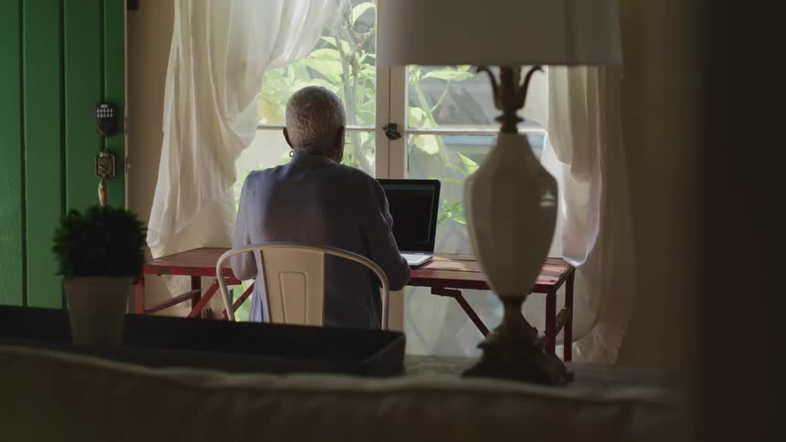 An elderly woman sits in her home and looks at her laptop