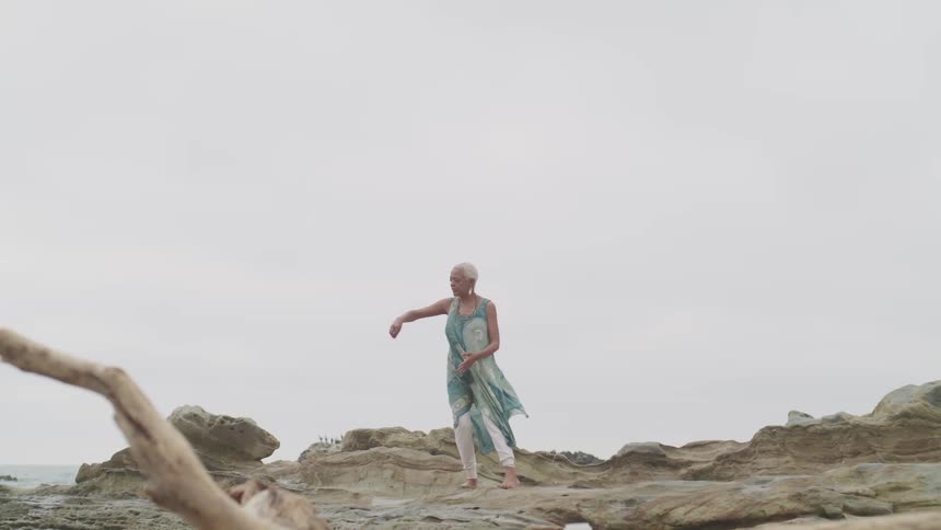 An elderly woman does yoga on a rock formation next to the ocean on a cloudy day