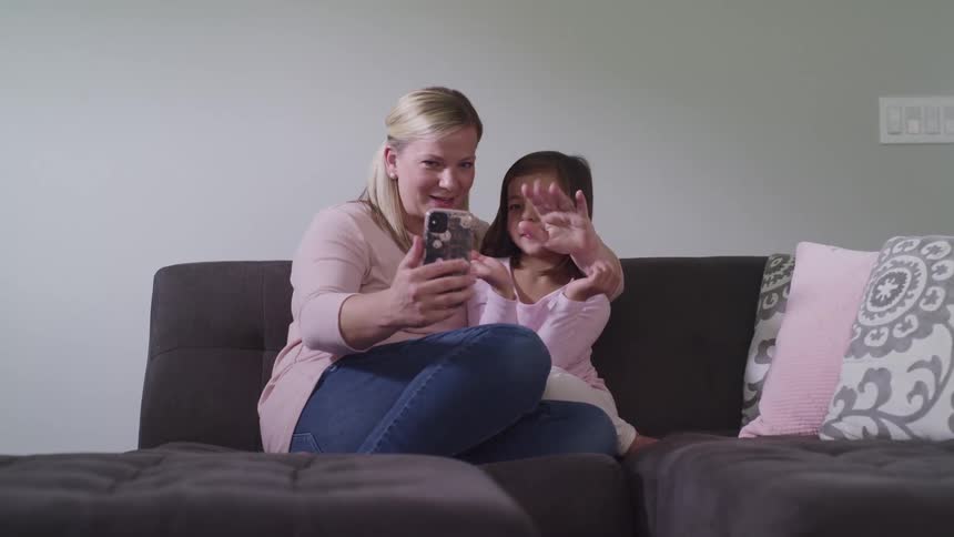 A mother and daughter sit on a couch and talk at wave at someone on a cell phone