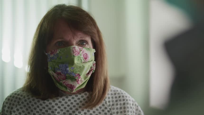 A woman with a mask and a hospital gown in a hospital with a window behind her