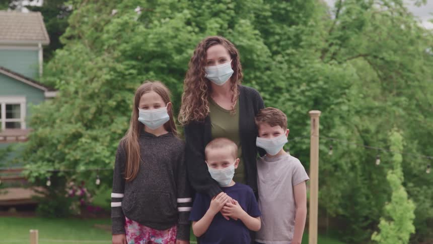 A woman a girl and two boys standing wearing masks with trees and a house in the background