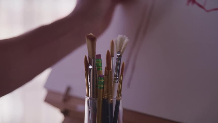 Glass filled with pencils and brushes in front of a person painting on a canvas over an easel at home