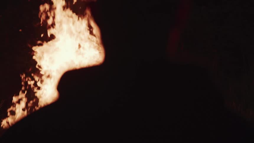 Man with hoodie is silhouetted against a fire that he watches at night