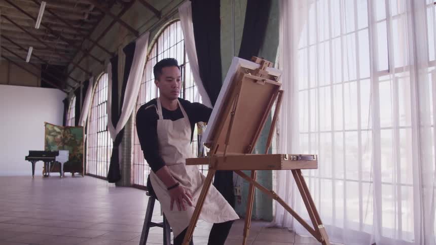 An asian man in a white apron sits on a stool in front of a canvas as he paints