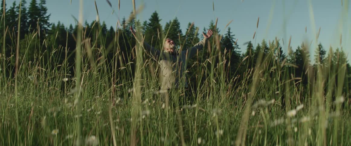 Senior stands in a grass field near pine trees with arms wide open at morning