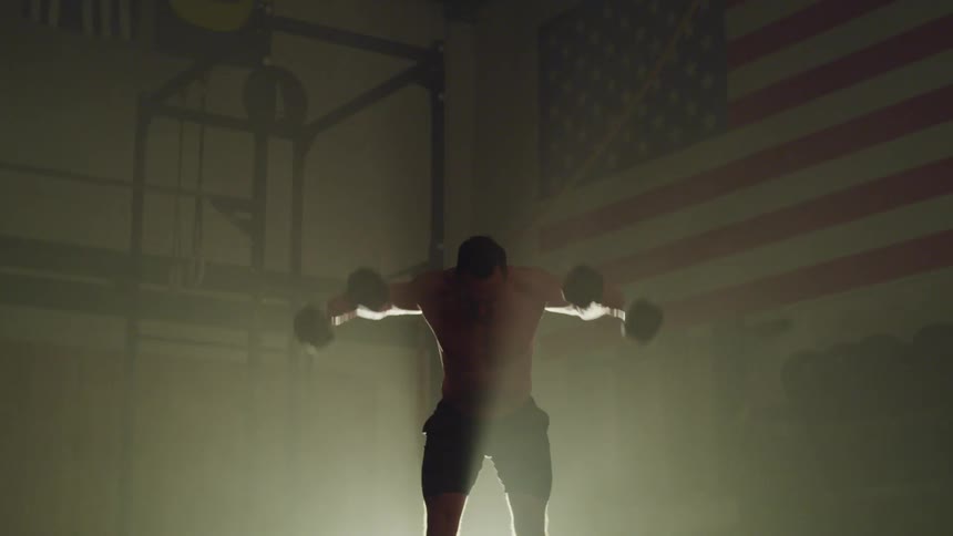 A man lifting dumbbells in a large gymnasium illuminated only by a spotlight
