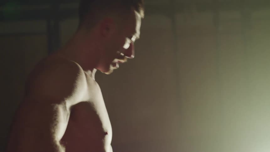 Bare chested man stretches and rubs his face standing in light cast by spotlight in dim room