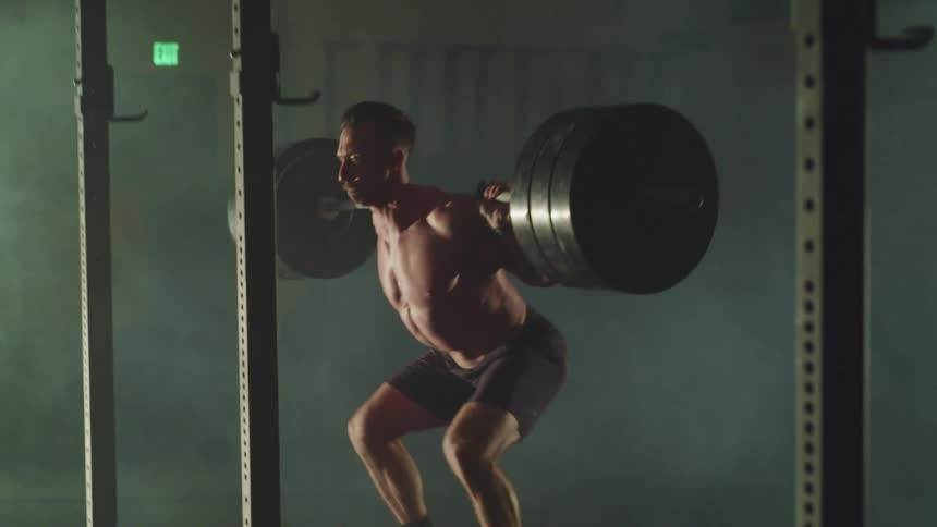 Shirtless strong muscular man deadlifts a loaded barbell on his shoulders in a dimly lit gym
