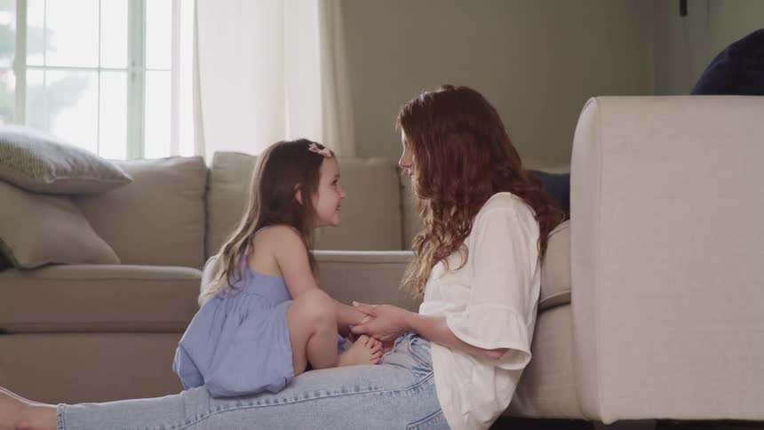 A mother sitting on the floor of her living room nuzzles her nose against her young daughters