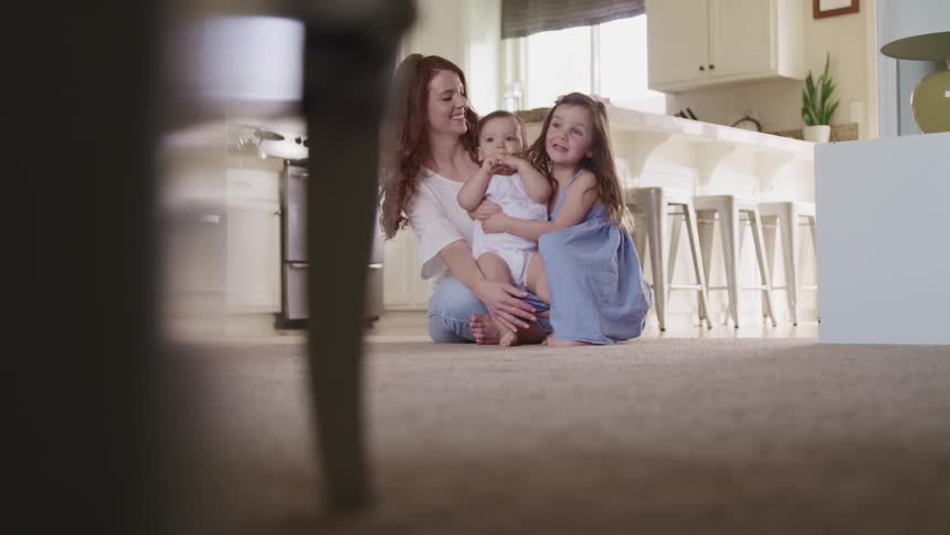 A woman sitting on the floor helps a baby to stand as a little girl hugs and kisses the baby