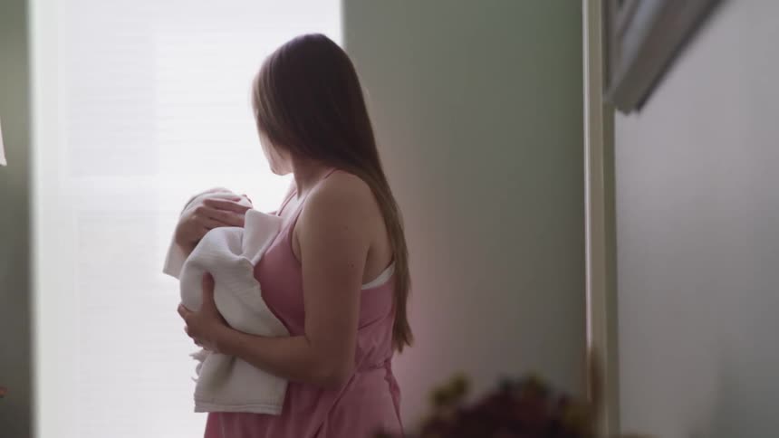 A woman stands by a window rocking a baby wrapped in a towel