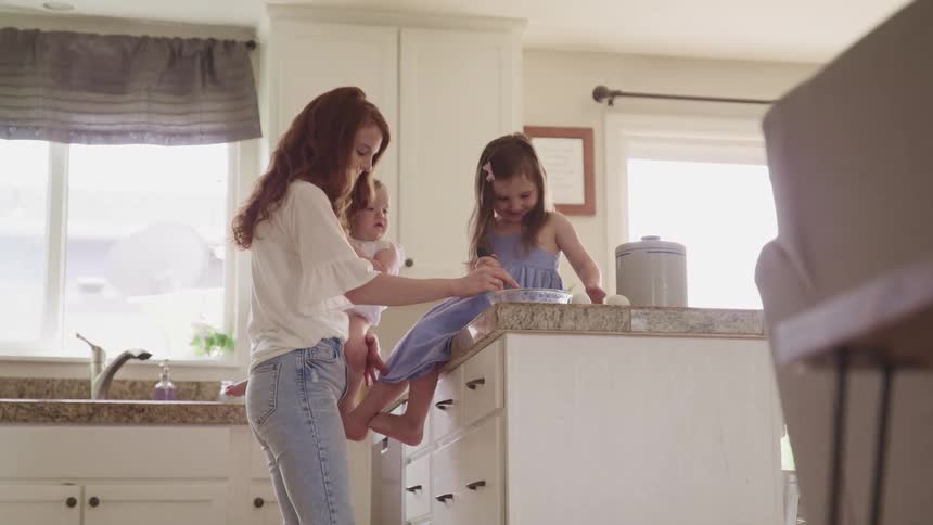 A woman holds a baby in the kitchen while a toddler girl sits on the kitchen counter