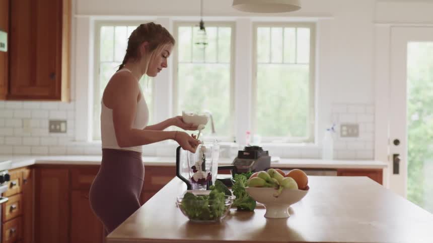 A young woman scoops cream into a blender filled with fruit