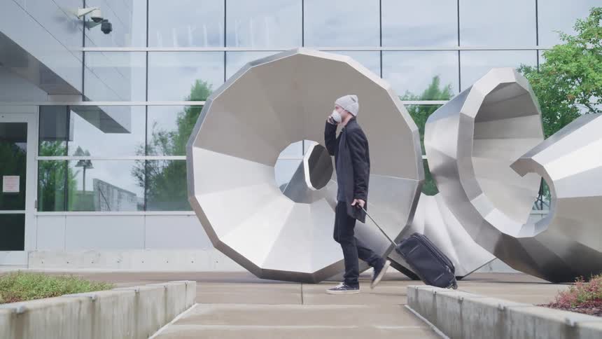 A man with a smart phone a mask and a hat pulling a luggage bag with a building in the background