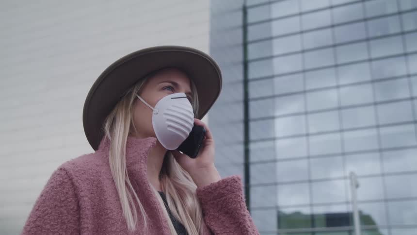 Woman wearing a mask holds a cell phone up to her ear while standing outdoors in a city