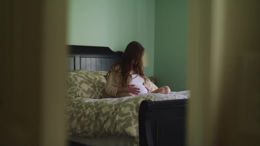 A pregnant woman sits on her bed while cradling her stomach