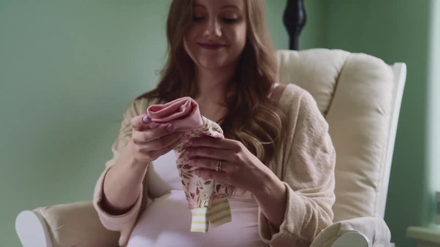 Pregnant woman with a long brown hair and sitting in a white armchair holds baby clothes and smiles warmly