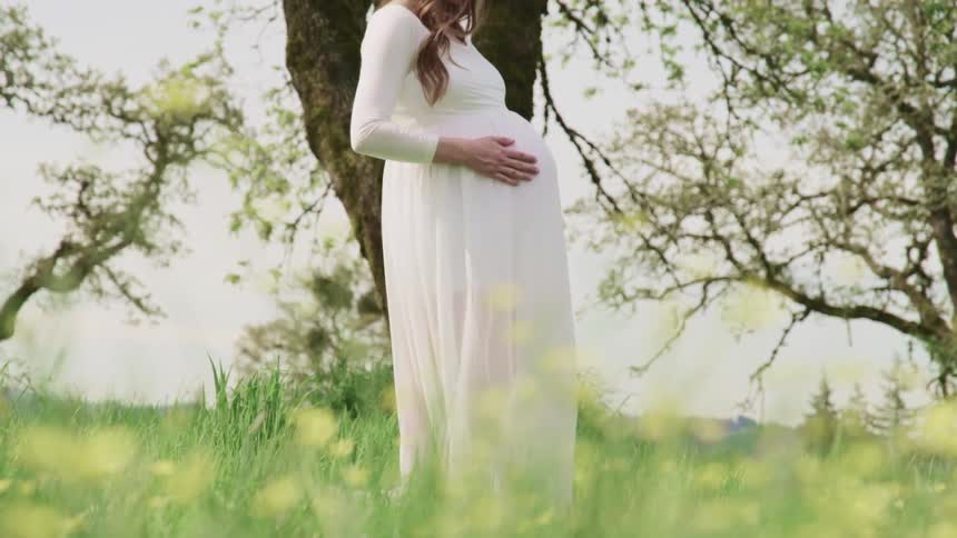 A pregnant woman strokes her stomach while standing outdoors in a field of grass
