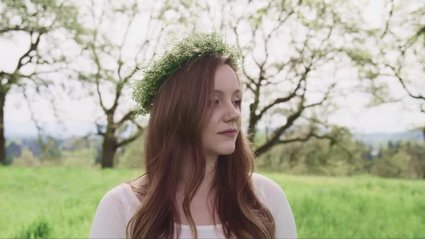 Side view of a pregnant woman with a white dress and a flower crown standing on grass with trees in the background
