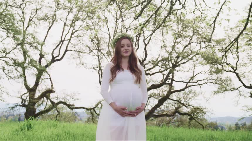 Pregnant woman in a long white dress and a flower crown or whreat on her head standing on a grass field