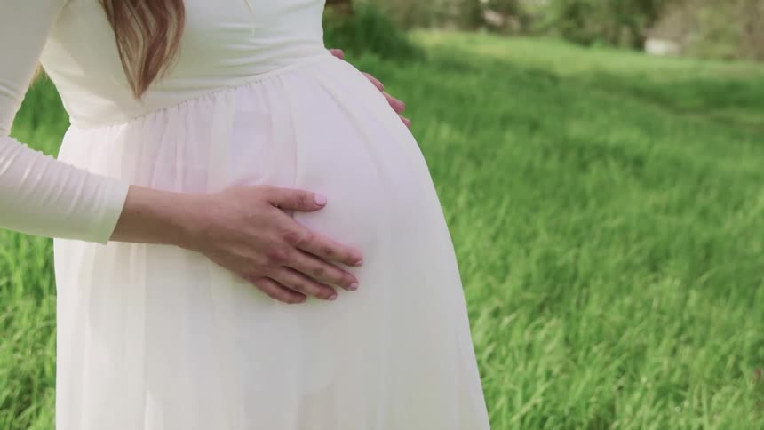 Woman with a white dress and a wedding ring standing on green grass holds her belly