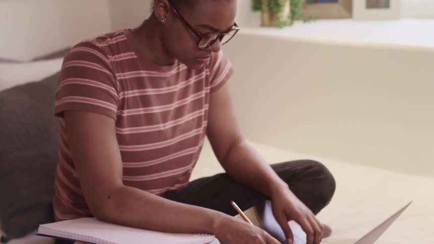 Woman sits in bed doing homework using a laptop and a notebook at home