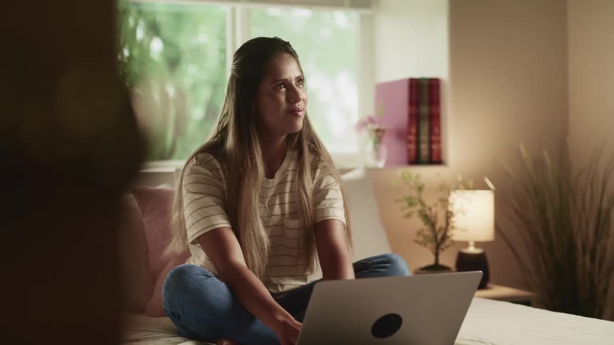 A teenager girl sits on her bed and types on a laptop