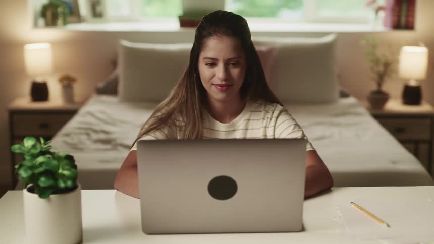 Brunette woman with brown long hair and a white t shirt sitting at a desk uses a laptop in her room