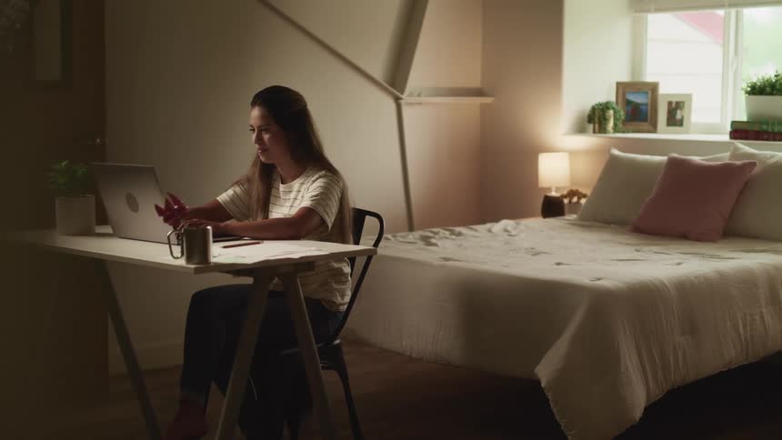 Brunette woman with long brown hair and a white shirt sits at a desk and uses a laptop in her room