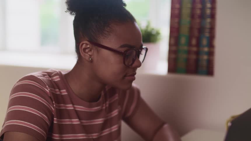 Black woman with glasses and striped t shirt sits at a desk and uses a laptop in a room