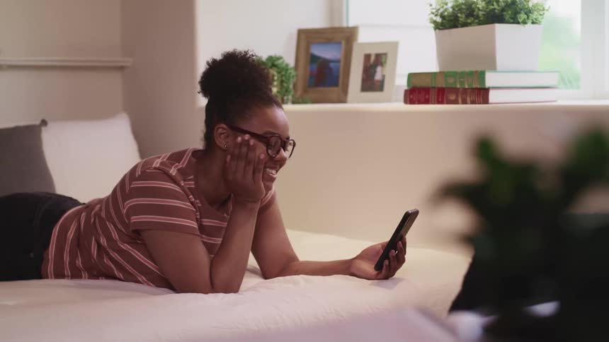 A young woman uses video chat on a smartphone while lying on her stomach on a bed