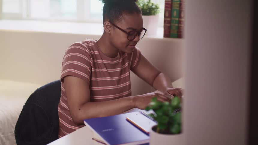Female smiles as she types on a laptop that is sitting on a desk in the bedroom along with a notebook and a pencil