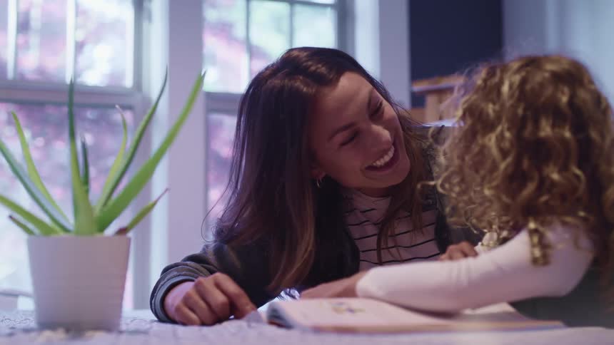 Mother and daughter smile at each other while reading a book in a table at home