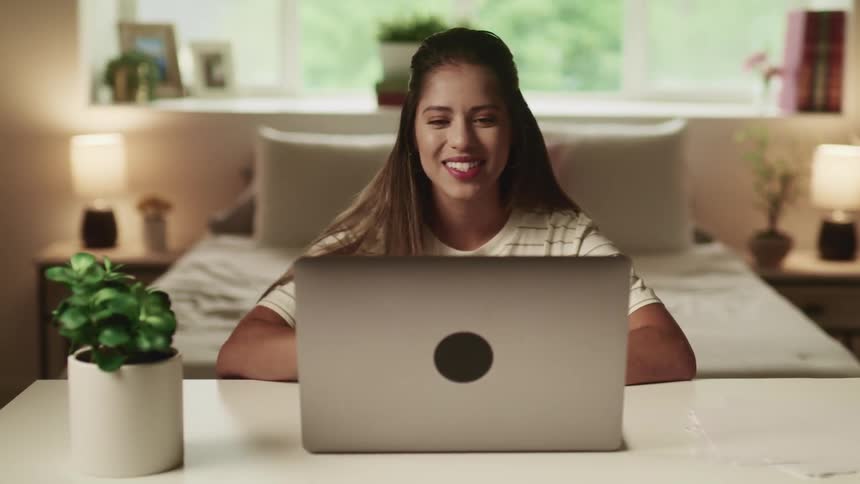 A female sits at a desk in her bedroom waving to a laptop