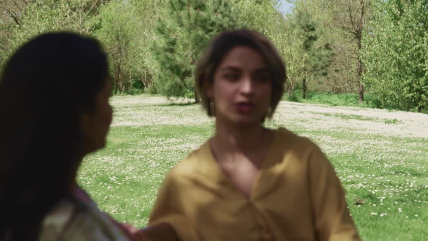 A pair of indian women talk with each other while sitting on a bench in a meadow