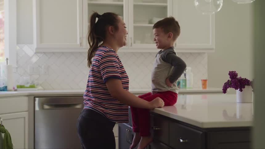 A woman makes faces as little boy sitting on the counter of a kitchen as the boy makes faces back