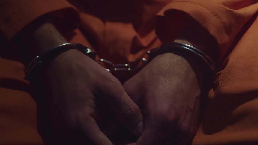 Close up of a man in an orange prison uniform with metallic handcuffs on his wrists