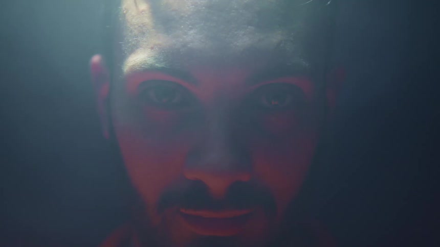 A man stands in a dark room under a swinging light and stares unblinking into the camera