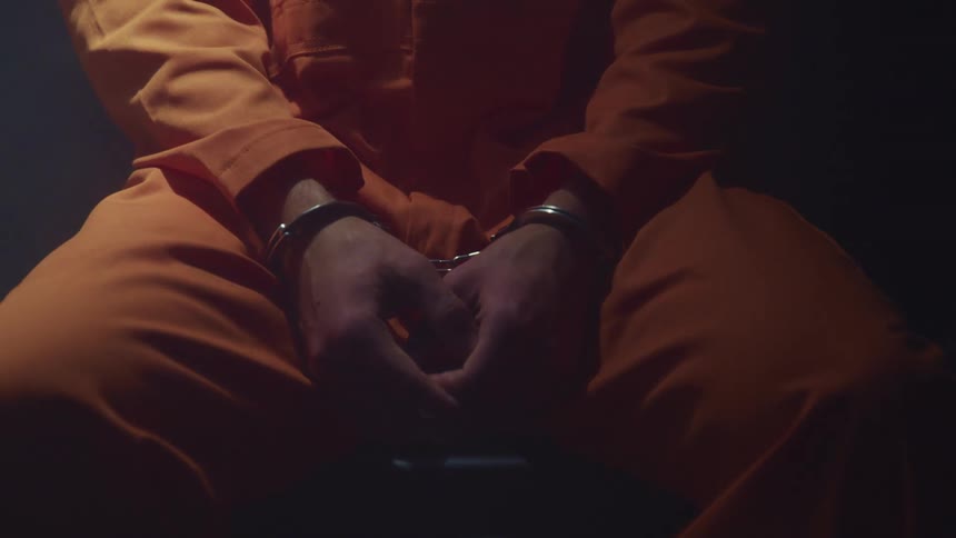 Man wearing an orange prison uniform and handcuffs sits in a dark room under a swinging white light