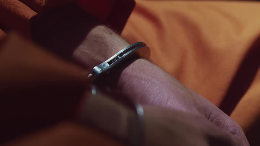 Person in inmate orange sits handcuffed as light and shadows change
