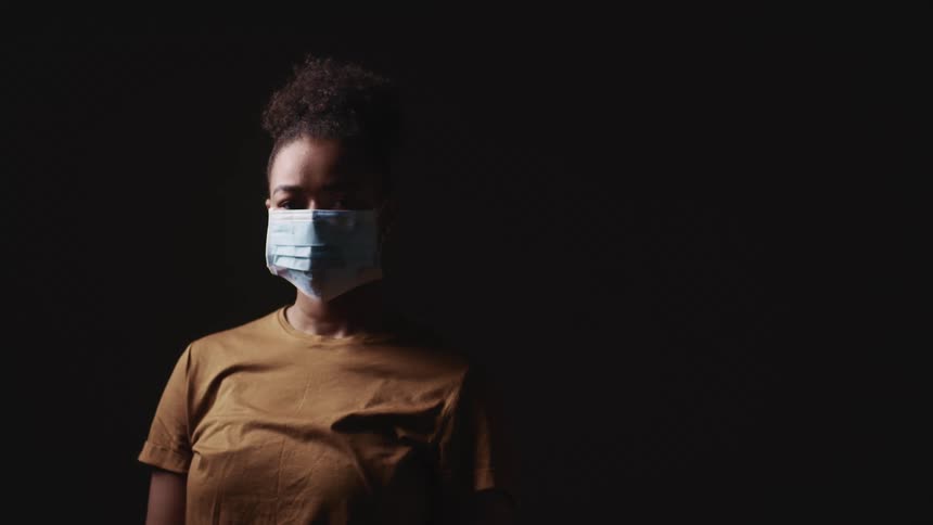 Black woman with a brown t shirt wears a face mask on the dark background
