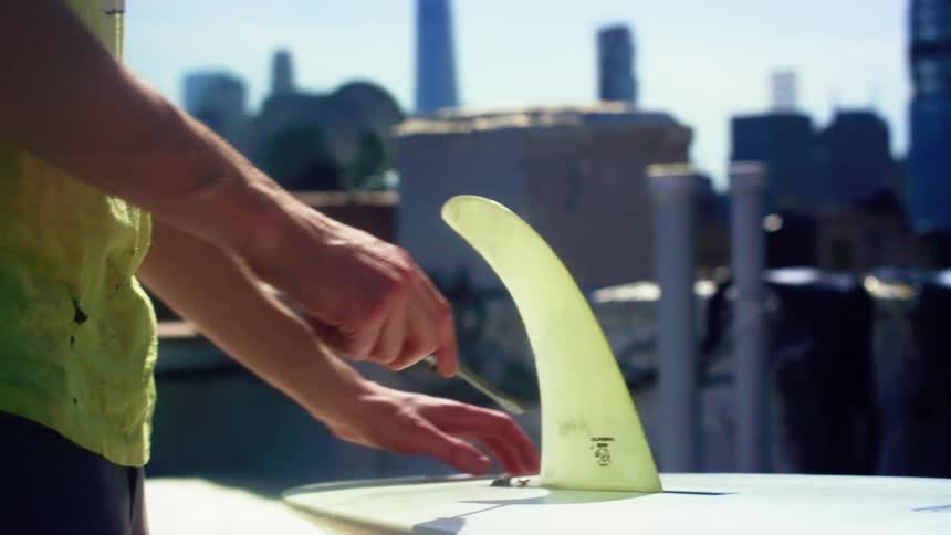 A man handing a screwdriver to another man who uses it to twist a screw on a surfboard