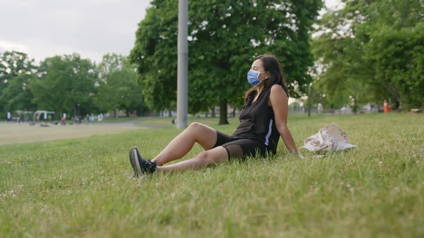 A woman in a medical mask relaxes in the grass near a sports field