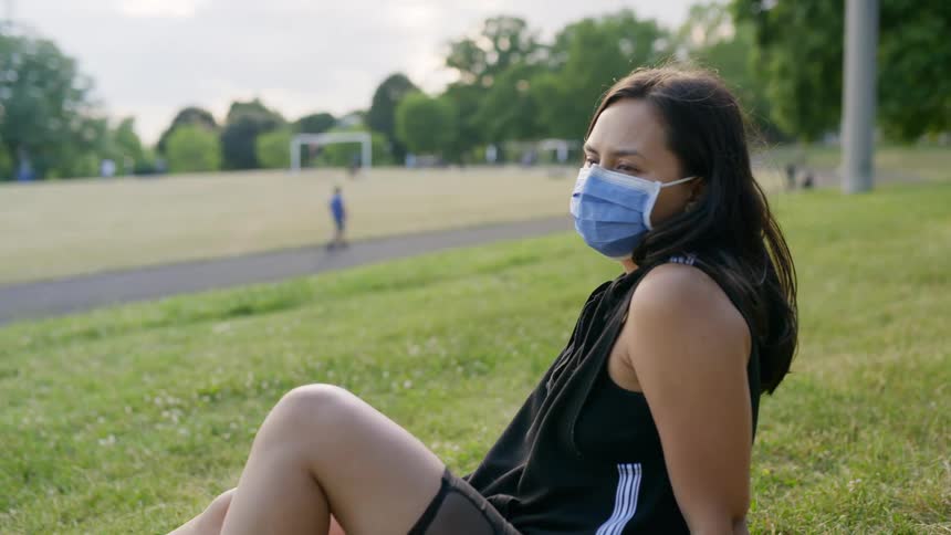 Woman wearing face mask relaxes on grass in park as other walk