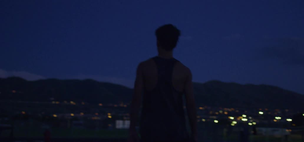 A teenager in running clothes walks toward the edge of a cliff overlooking a neighborhood by a mountain range