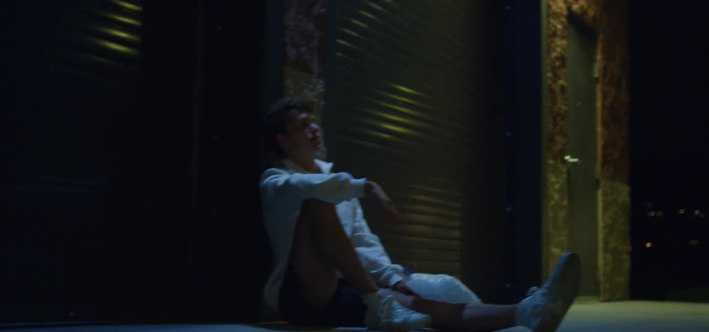 A young man sits on the ground with his back against the wall of a garage outdoors at night